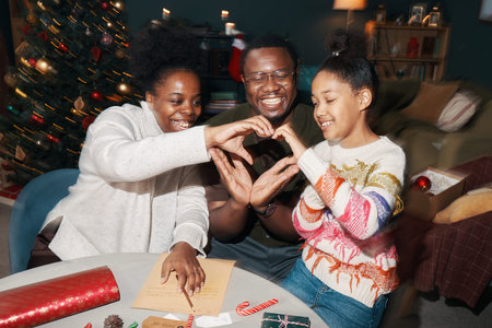 Black woman, Black man, and Black girl sitting together making heart shape with hands, smiling and...