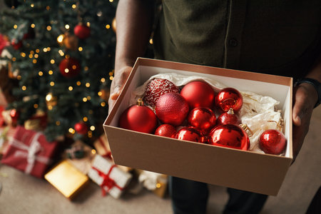 Person holding box filled with red Christmas ornaments near decorated Christmas tree with wrapped gifts underneath, hands visible presenting festive decorations for holiday preparationの写真素材