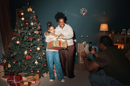 Woman and girl standing beside decorated Christmas tree holding wrapped gifts while man, kneeling in front taking photo with smartphone in cozy living roomの写真素材