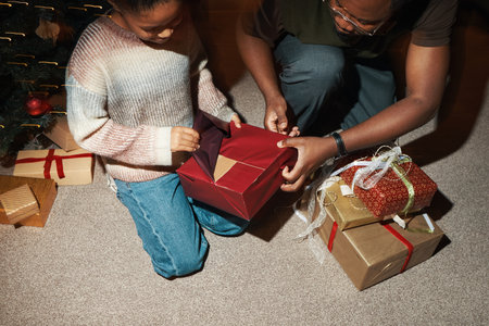 Child opening gift box with man assisting beside Christmas tree, several wrapped presents stacked on carpet, both focused on unwrapping holiday present togetherの写真素材