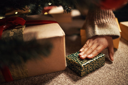 Hand of young child reaching for wrapped Christmas gift under tree, fingers touching small present with festive pattern, surrounded by other boxes, holiday celebration sceneの写真素材