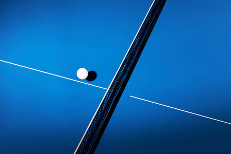 Closeup showing ping pong ball resting on blue table tennis surface with net and lines visible, capturing minimalistic sports scene with strong geometric compositionの写真素材