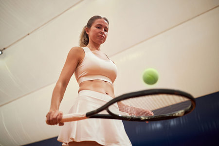 Caucasian young adult woman playing tennis indoors, holding racket and preparing to hit tennis ball, focused expression, athletic stance, visible sportswear, indoor court backgroundの写真素材