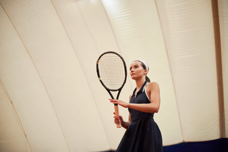 Caucasian young adult woman holding tennis racket preparing to serve during indoor match, focused expression, athletic stance, headband visible, looking forwardの写真素材