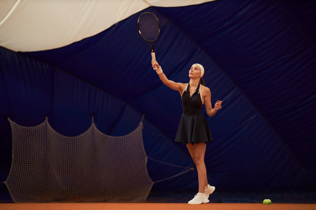 Caucasian young adult woman preparing to serve tennis ball on indoor court, holding racket overhead and focusing on ball, wearing athletic dress and headband, net and ball visibleの写真素材