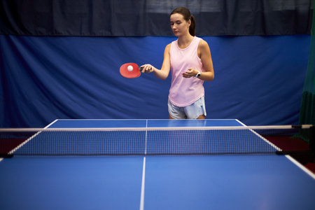 Young adult Caucasian woman practicing table tennis, holding paddle in right hand and tossing ball with left hand, standing on one side of blue ping pong table indoorsの写真素材