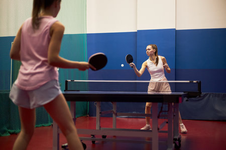 Two Caucasian teenage girls playing table tennis indoors, both holding paddles and focusing on ball, standing on opposite sides of table in sports facility with blue and red flooringの写真素材