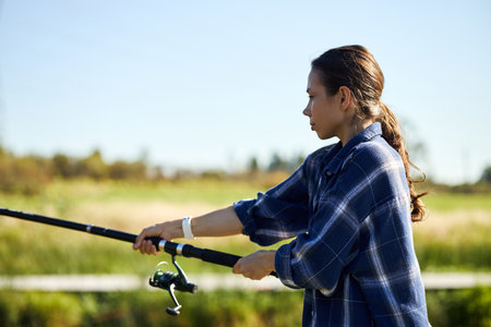 Young adult woman fishing outdoors, holding fishing rod with both hands, standing in profile against blurred green landscape, focused on casting line, long hair tied backの写真素材