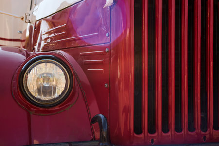 Closeup showing front section of vintage red truck with round headlight and vertical grille, capturing classic automotive design details and reflecting sunlight on metal surfaceの写真素材
