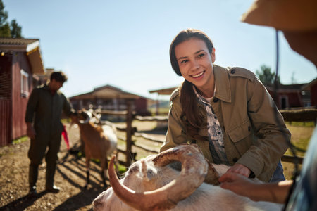 Caucasian young adult woman smiling while petting goat outdoors on farm, interacting with animal as two people stand in background near barn structures under sunny skyの写真素材