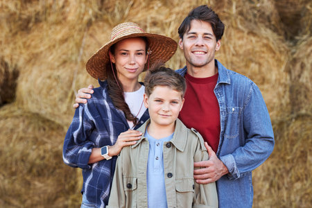 Caucasian young adult woman wearing straw hat standing beside Caucasian young adult man with hand on shoulder of Caucasian boy, all smiling and posing in front of hay balesの写真素材