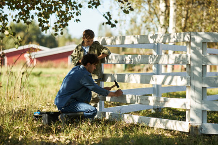 Caucasian middle aged man repairing wooden fence with hammer while Caucasian boy watching outdoors on sunny day, rural setting with trees and grass in background, teamwork conceptの写真素材