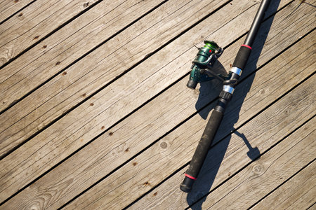 Fishing rod lying diagonally on wooden dock, casting strong shadow across planks, closeup showing reel and handle, outdoor recreational equipment resting on sunlit surfaceの写真素材