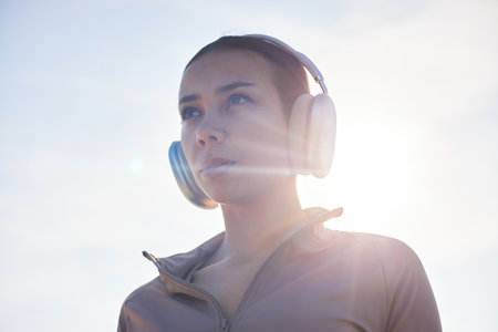 Portrait of young adult Asian woman wearing headphones gazing into distance outdoors, sunlight creating lens flare effect, subject appearing focused and contemplativeの写真素材