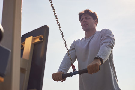 Caucasian young adult man exercising on outdoor fitness equipment gripping handles and pulling chain looking focused during workout session under clear skyの写真素材