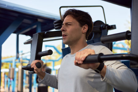 Young adult man exercising on outdoor gym machine, gripping handles and focusing ahead, wearing...