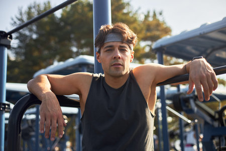 Portrait of young adult Caucasian man resting after workout outdoors, leaning on exercise equipment,...