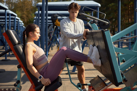 Young adult woman exercising on leg press machine outdoors while young adult man supervising...