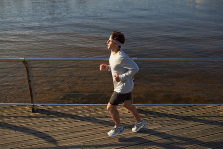 Young adult man jogging along waterfront boardwalk, maintaining steady pace with focused expression,...