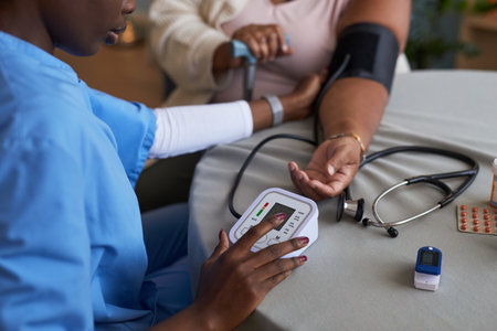 Black female nurse measuring blood pressure of geriatric female patient using digital monitor, stethoscope and pulse oximeter on table, arm extended for examinationの写真素材