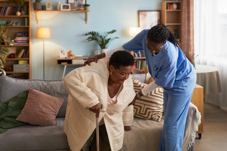 Senior Black woman using cane receiving assistance from young adult Black female nurse in scrubs, helping her stand up from sofa in living room setting, during home care visitの写真素材