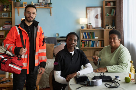 Caucasian young adult man and Black young adult woman paramedics assisting middle aged Black woman measuring blood pressure at home, medical equipment and supplies on tableの写真素材