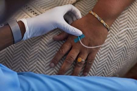 Middle aged Black woman receiving intravenous therapy in hand while healthcare worker wearing glove assisting, close up of hands showing medical procedure and patient jewelryの写真素材