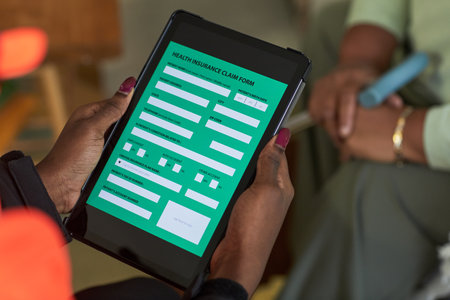 Black female nurse holding digital tablet displaying patient health insurance claim form, sitting indoors with another in background, hands visible interacting with electronic deviceの写真素材