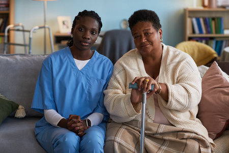 Portrait of young adult Black woman caregiver sitting beside senior Black woman with disability holding cane, both looking at camera, showing support and companionship on sofa during home visitの写真素材