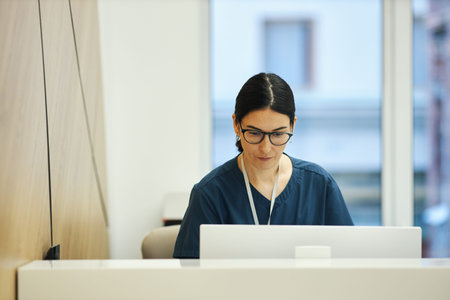 Caucasian young female nurse wearing glasses working on laptop at reception desk in medical office at clinic, focused on screen, dark hair tied back, professional environment visibleの写真素材