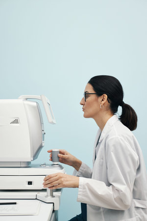 Young Caucasian female doctor operating ophthalmic diagnostic machine in medical clinic, holding...
