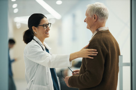 Caucasian female doctor greeting senior Caucasian male patient in medical facility, doctor smiling and placing hand on patients shoulder, while holding eyeglasses in other handの写真素材