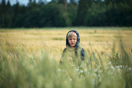 Caucasian child standing in tall grass field, looking forward with neutral expression, surrounded by wild vegetation and blurred background of trees and open landscapeの写真素材