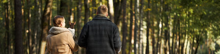 Header of Caucasian middle aged man and Caucasian young adult woman walking together in forest holding small dog, both seen from behind, surrounded by tall trees in natural settingの写真素材