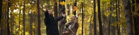 Header of Caucasian young adult man and Caucasian young adult woman standing in forest throwing autumn leaves upward, smiling and looking at each other surrounded by tall treesの写真素材