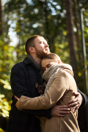 Caucasian young adult man embracing Caucasian young adult woman outdoors in forest, woman resting head on mans chest while holding small dog, both appearing relaxed and affectionateの写真素材