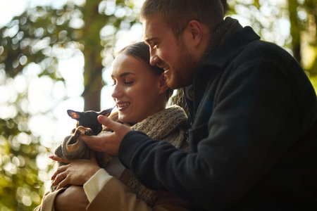 Caucasian young woman and man holding small chihuahua dog outdoors, smiling and interacting with pet, standing close together in natural forest setting, enjoying leisure timeの写真素材