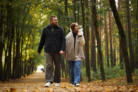 Caucasian young adult man and Caucasian young adult woman walking together holding hands in forest park, both looking at each other and smiling, autumn leaves covering groundの写真素材
