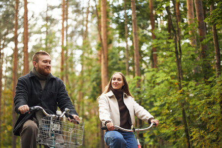 Young adult man and young adult woman riding bicycles through forest, both smiling and looking at each other, surrounded by tall trees and greeneryの写真素材