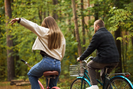 Caucasian young woman riding bicycle with arm raised next to Caucasian young man cycling through forest, both seen from behind, enjoying outdoor activity in wooded areaの写真素材