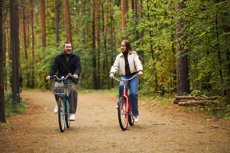 Caucasian young adult man and Caucasian young adult woman riding bicycles together on forest path,...