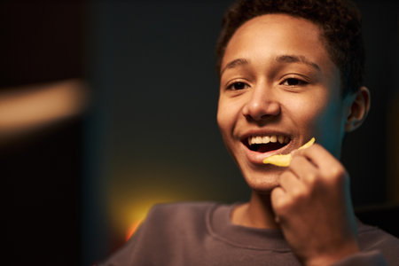 Portrait of teenage Black boy smiling and holding potato chip near mouth, looking slightly off camera, showing joyful expression while enjoying snack in casual indoor settingの写真素材