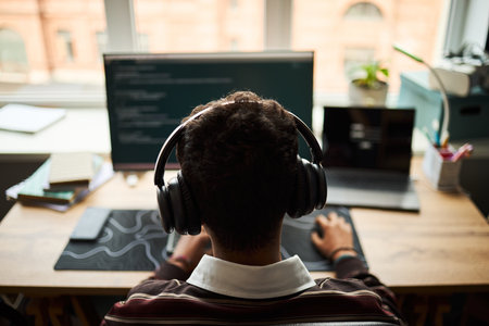 Young adult man sitting at desk wearing headphones working on computer with code on monitor, laptop open beside him, focused on programming in modern office settingの写真素材