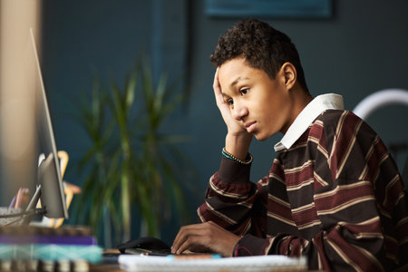 Teenage boy sitting at desk using desktop computer, resting head on hand, appearing focused while studying or working on assignment in modern indoor settingの写真素材