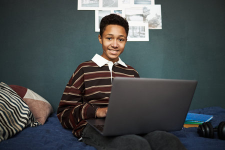 Portrait of Black teenager boy sitting on bed using laptop, smiling at camera, working or studying...