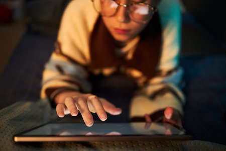Caucasian child wearing glasses using digital tablet while lying on bed, focusing on screen with hand touching device, demonstrating technology interaction in home environmentの写真素材