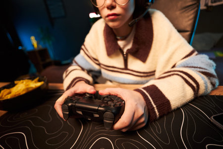 Teenager girl wearing headset holding game controller sitting at desk playing video game, hands...