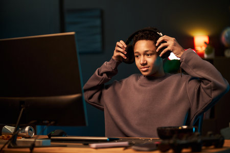Black teenage boy sitting at desk putting on headphones while looking at computer monitor, engaging in gaming or online activity in modern room with electronic devices visibleの写真素材