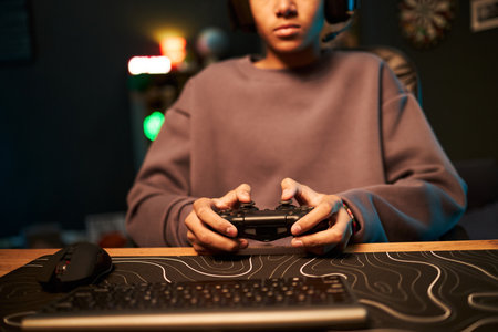 Teenage boy with medium skin tone wearing headset sitting at desk holding game controller playing video game, keyboard and mouse visible in foreground, focused on screenの写真素材
