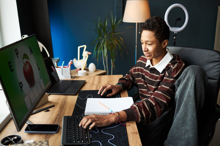 Teenage Black boy sitting at desk using computer mouse and keyboard while looking at monitor,...